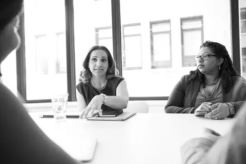 Black-and-white photo of two women at a table in a workplace; one is addressing people outside the frame, while the other listens.